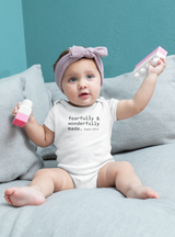 A baby girl wearing a white fearfully and wonderfully made onesie sits on a grey couch holding pink and white building blocks.