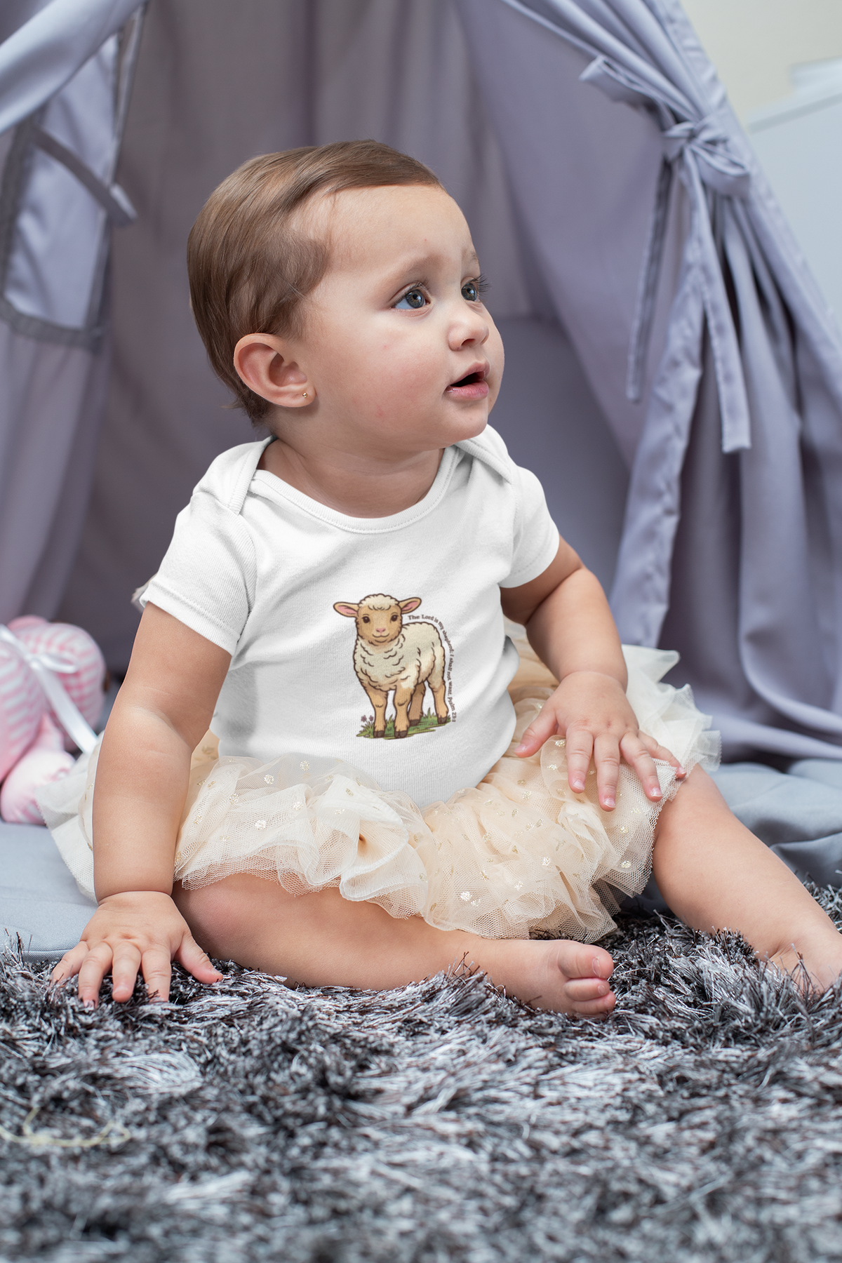 A baby girl wearing a white Psalm 23 baby onesie and a beige tutu sits gracefully on a fluffy grey rug in front of a grey play tent.