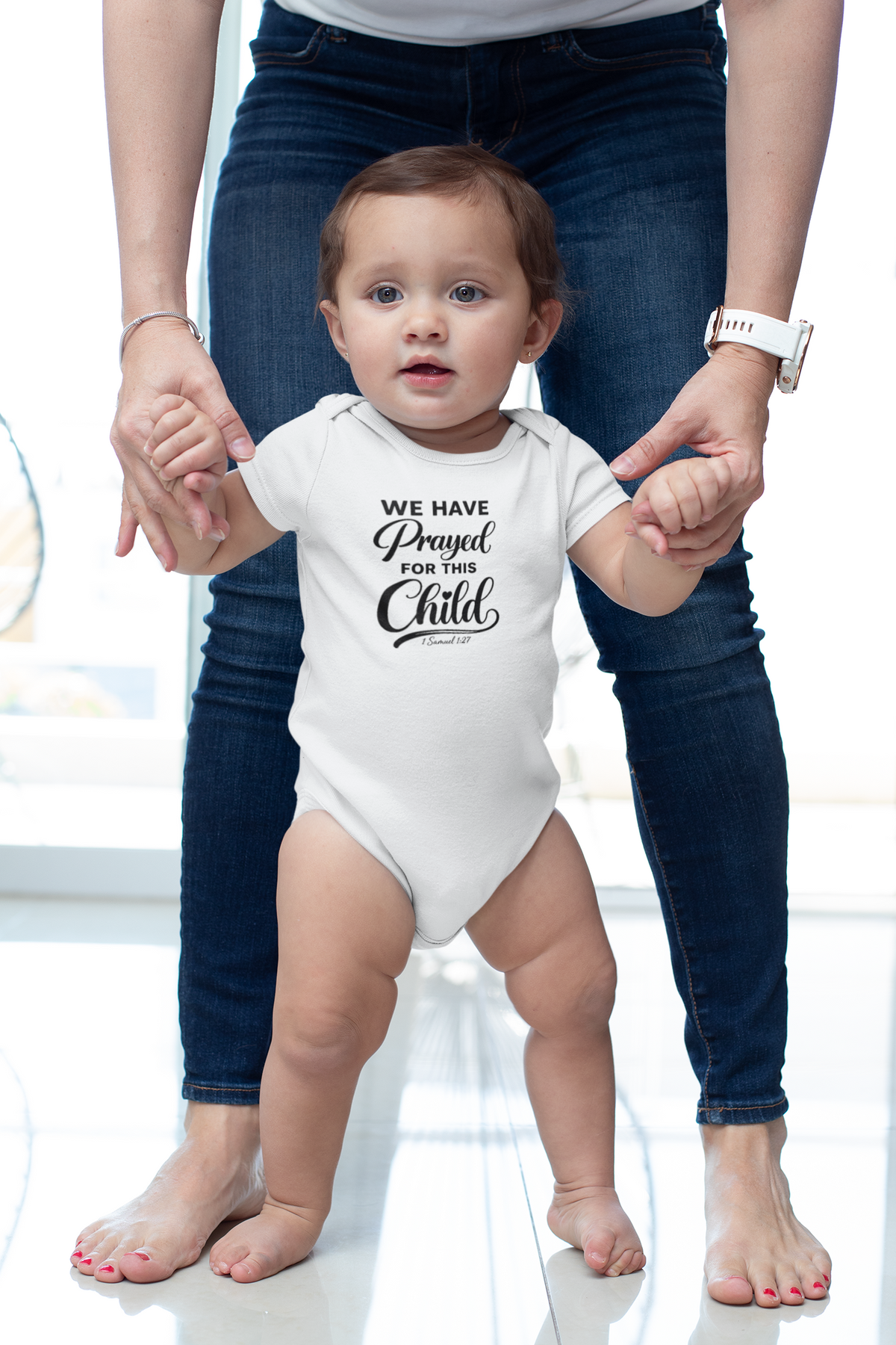A toddler wearing a white We Have Prayed For This Child onesie takes steps while holding the hands of an adult standing behind them in a bright room.