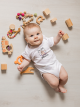 A happy baby wearing a white Our Little Miracle bodysuit lies on a light wooden floor surrounded by wooden toys and a pacifier clip.