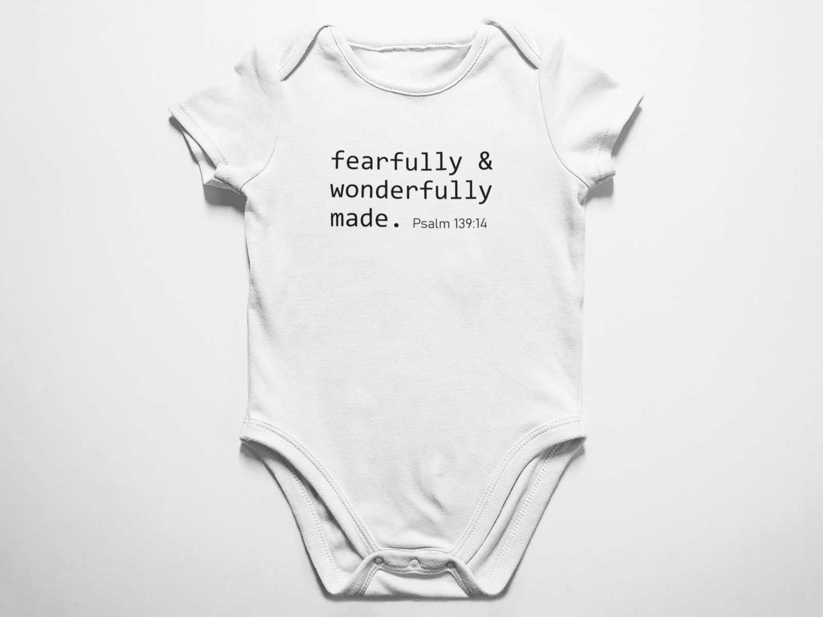 A clean flat lay view of a white baby onesie featuring the black typewriter text fearfully and wonderfully made Psalm 139 verse 14 against a white background.