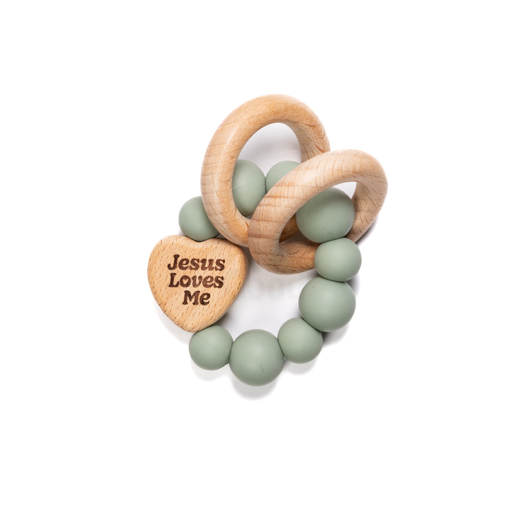 Baby teething ring with sage green silicone beads, wooden rings, and engraved Jesus Loves Me heart photographed on white.