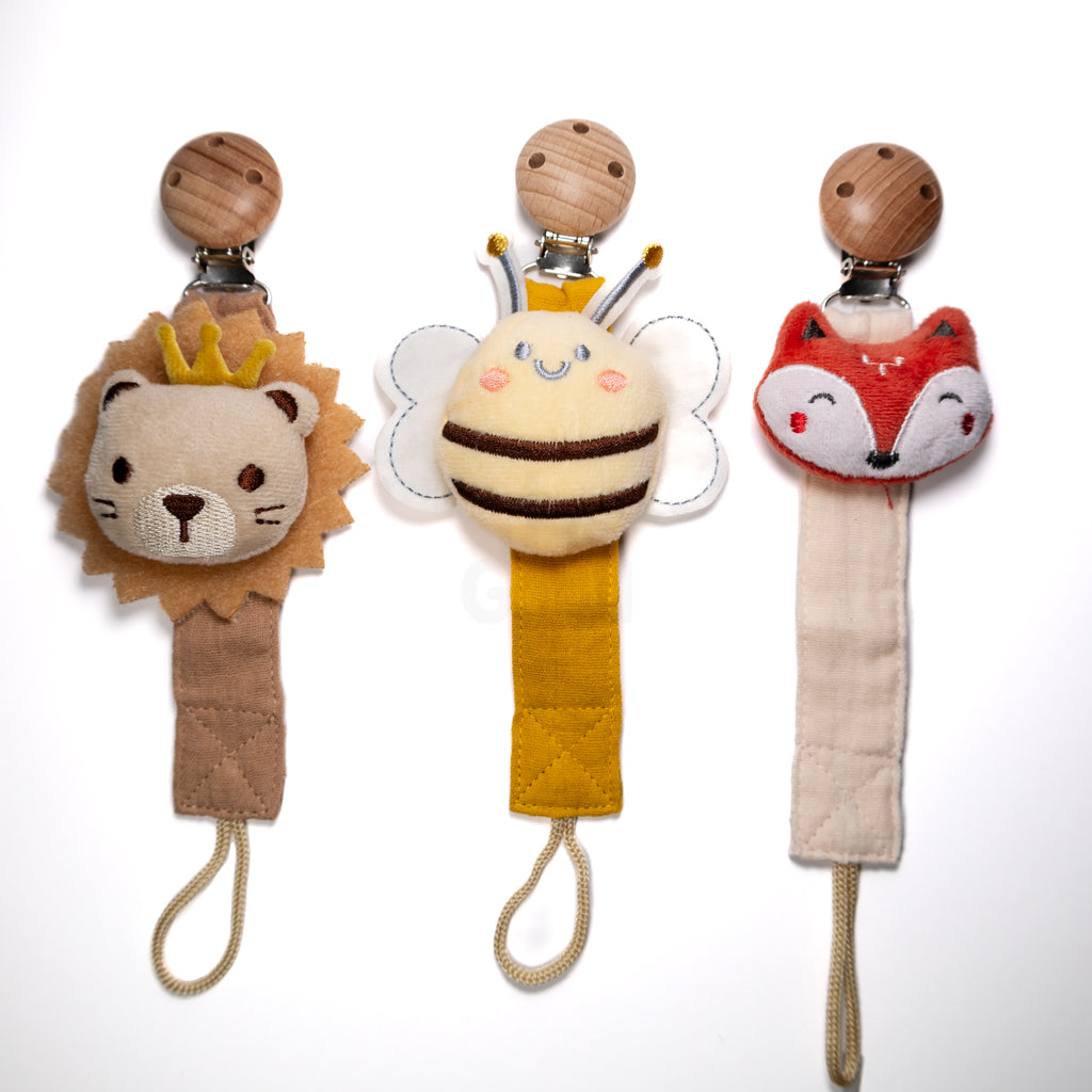Three baby pacifier clips displayed side by side featuring a plush lion with a crown, a smiling bee with wings, and a fox face, each with a fabric strap and wooden clip on a white background, sold as a combo pack for babies and toddlers