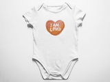 A clean flat lay view of a white baby onesie featuring a warm watercolor heart and the white text I am loved against a white background.