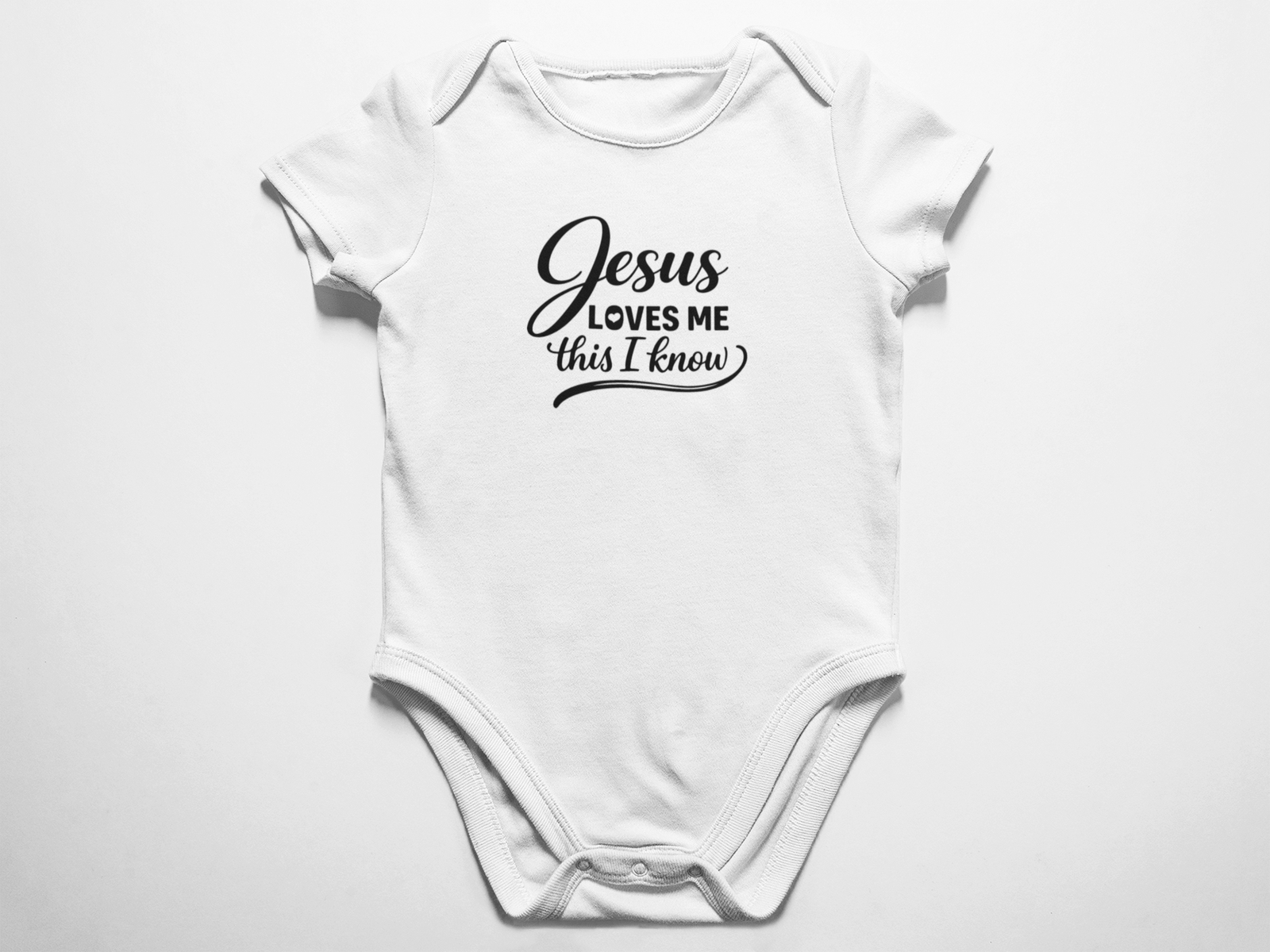 A clean flat lay view of a white baby onesie featuring the black text Jesus Loves Me This I know in a mix of cursive and classic typography against a white background.
