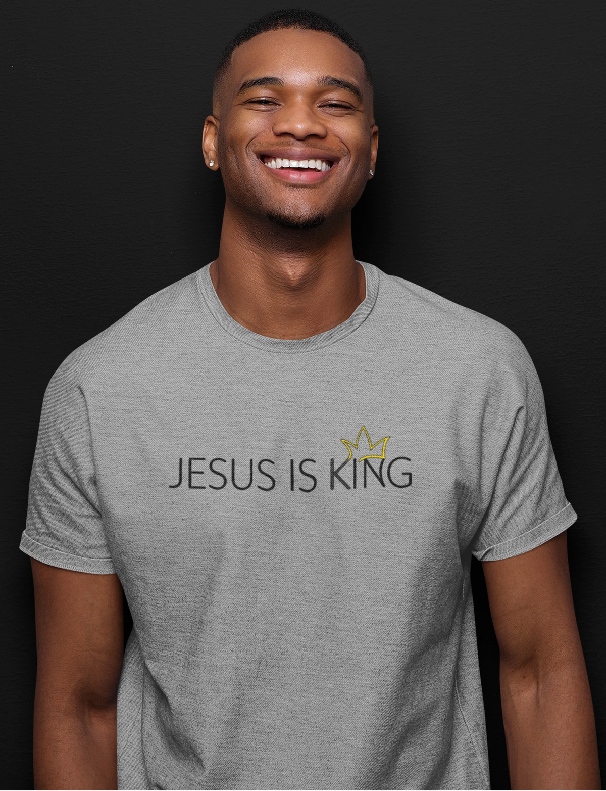 A smiling man wearing a heather gray t-shirt with the text 'JESUS IS KING' and a small gold crown graphic on the chest, set against a solid black background.