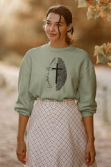 A woman wearing a sage green crewneck sweatshirt with a black graphic of a cross, an eye of Providence, and the words 'Created in the image,' paired with a pink and white checkered skirt, set against a warm, blurred outdoor background.