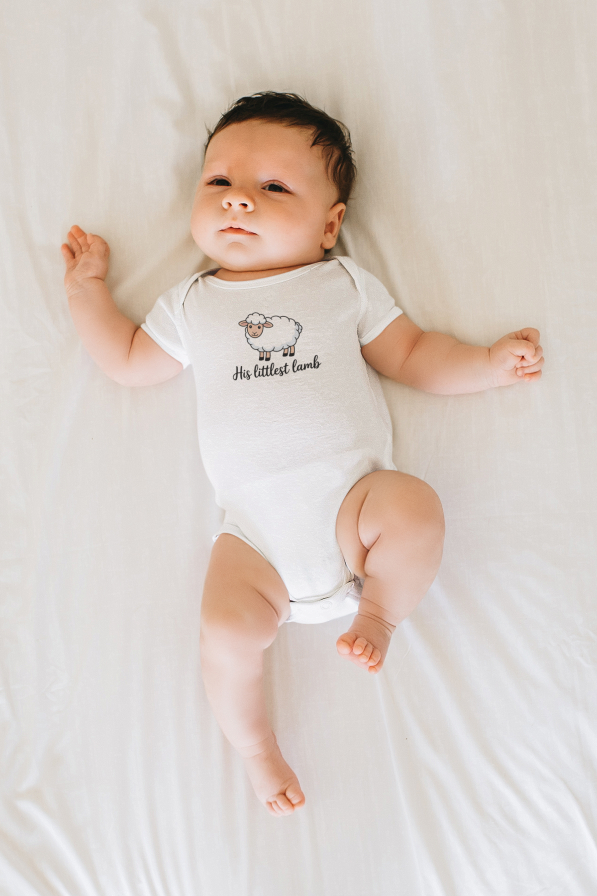 A sweet newborn baby wearing a white His littlest lamb onesie lies peacefully on a soft white bed sheet.