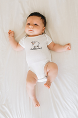 A sweet newborn baby wearing a white His littlest lamb onesie lies peacefully on a soft white bed sheet.