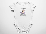A clean flat lay view of a white baby bodysuit featuring the text Our Little Miracle with an illustration of praying hands surrounded by pastel hearts against a white background.