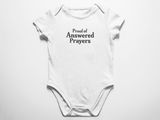 A clean flat lay view of a white baby onesie featuring the text Proof of Answered Prayers in bold classic typography against a white background.