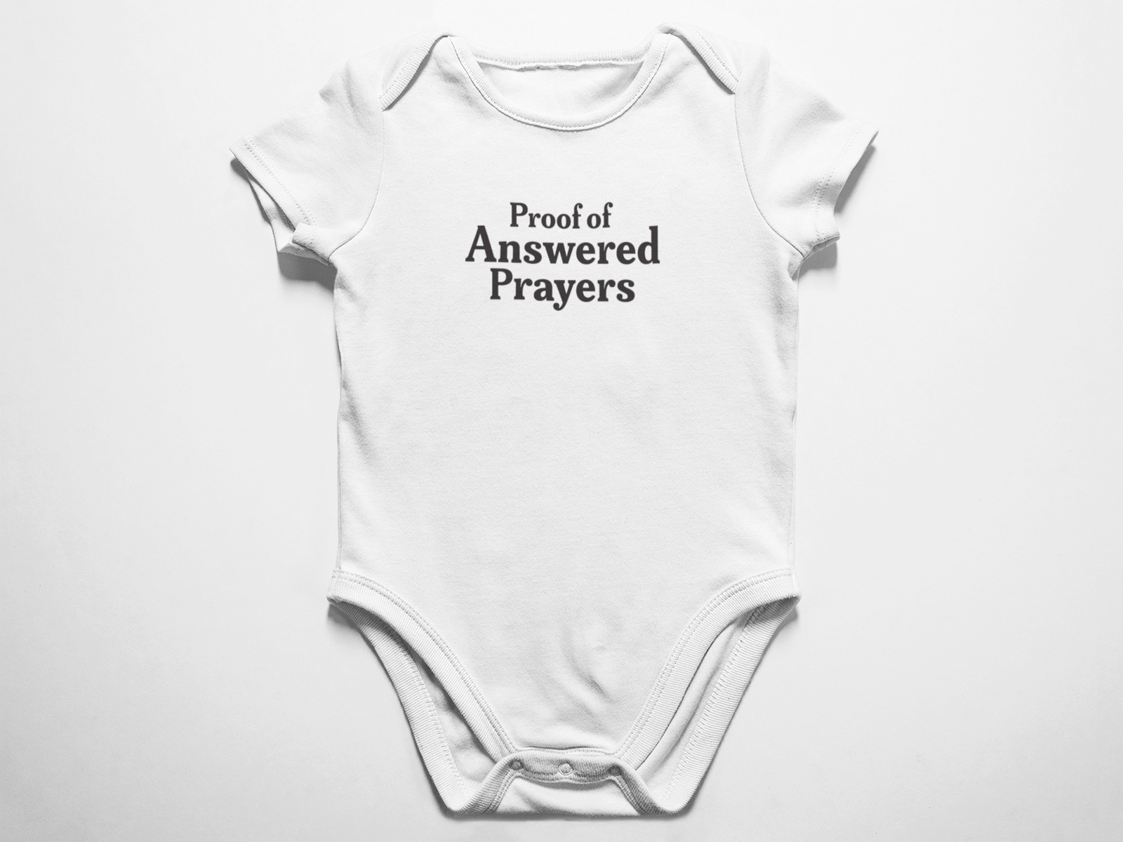A clean flat lay view of a white baby onesie featuring the text Proof of Answered Prayers in bold classic typography against a white background.