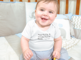 A smiling baby wearing a white fearfully and wonderfully made onesie sits happily in a crib with blue and grey chevron bedding.