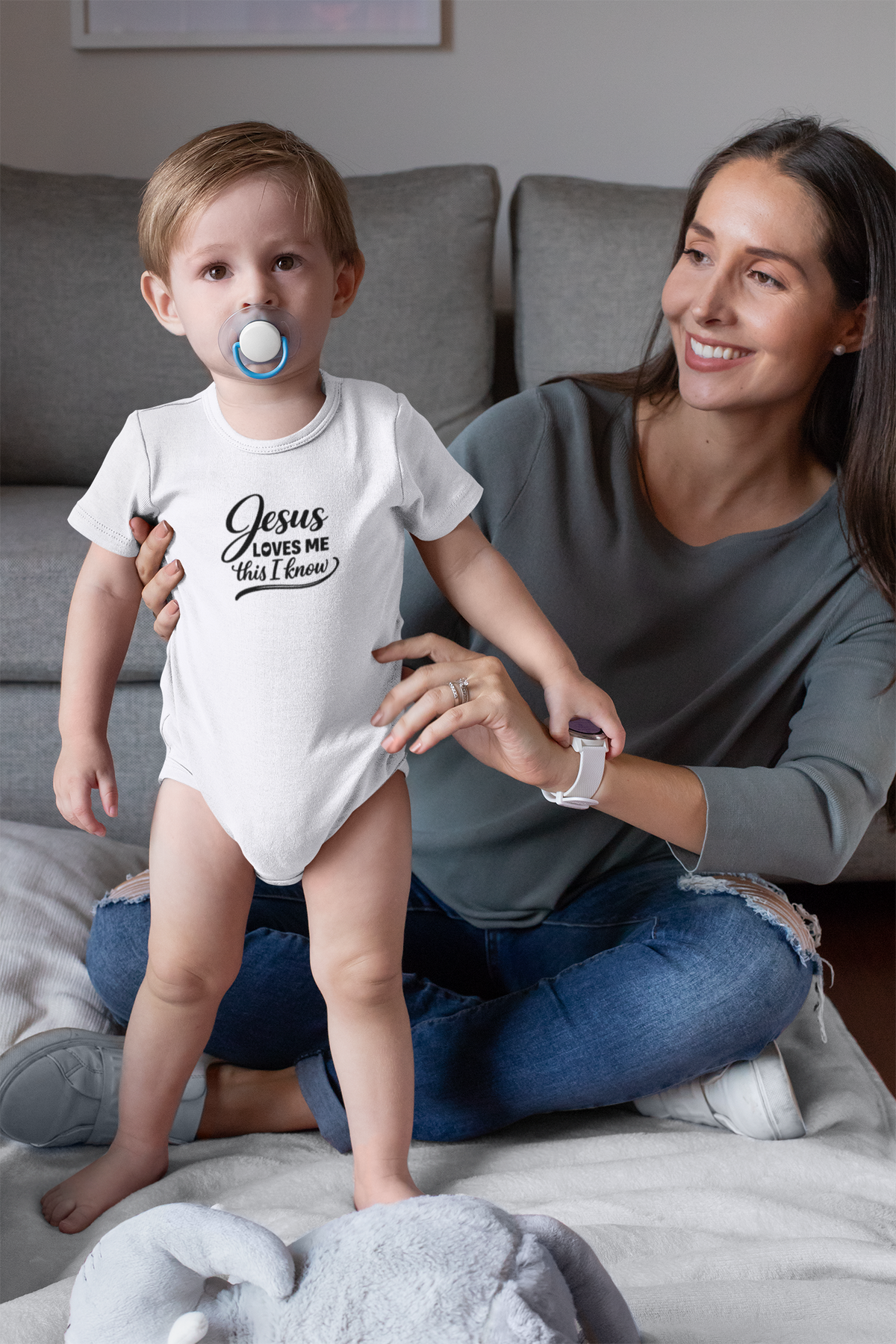 A toddler wearing a white baby onesie with the text Jesus Loves Me This I know stands playfully while a smiling mother sits on the floor beside him in a cozy living room.