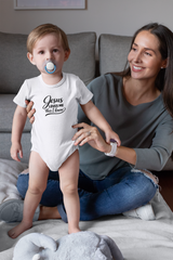 A toddler wearing a white baby onesie with the text Jesus Loves Me This I know stands playfully while a smiling mother sits on the floor beside him in a cozy living room.