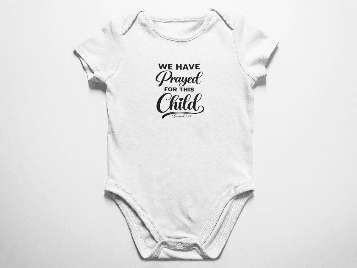A clean flat lay view of a white baby onesie featuring the black text We Have Prayed For This Child and 1 Samuel 1 verse 27 against a white background.