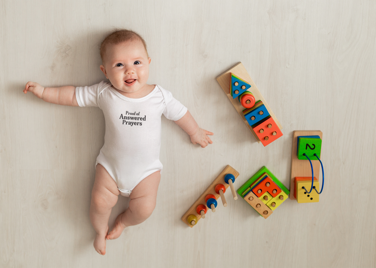 A smiling baby wearing a white Proof of Answered Prayers bodysuit lies on a light wooden floor surrounded by colorful wooden educational toys.