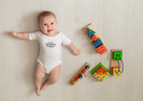 A smiling baby wearing a white Proof of Answered Prayers bodysuit lies on a light wooden floor surrounded by colorful wooden educational toys.